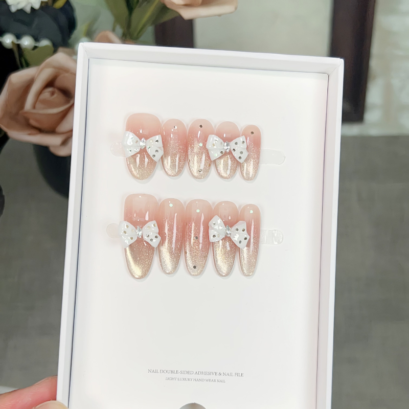 Kézzel készített körmök arany macskaszem effektussal, fehér masnival, gyengéd blush árnyalattal, nyári manikűr